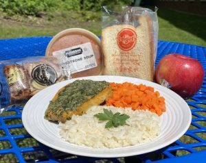 Yellow chicken patty with white rice and orange diced carrots on a white plate, with an apple, bread, minestrone soup in a container, and a marble cake dessert. All on a blue table with grass in the background